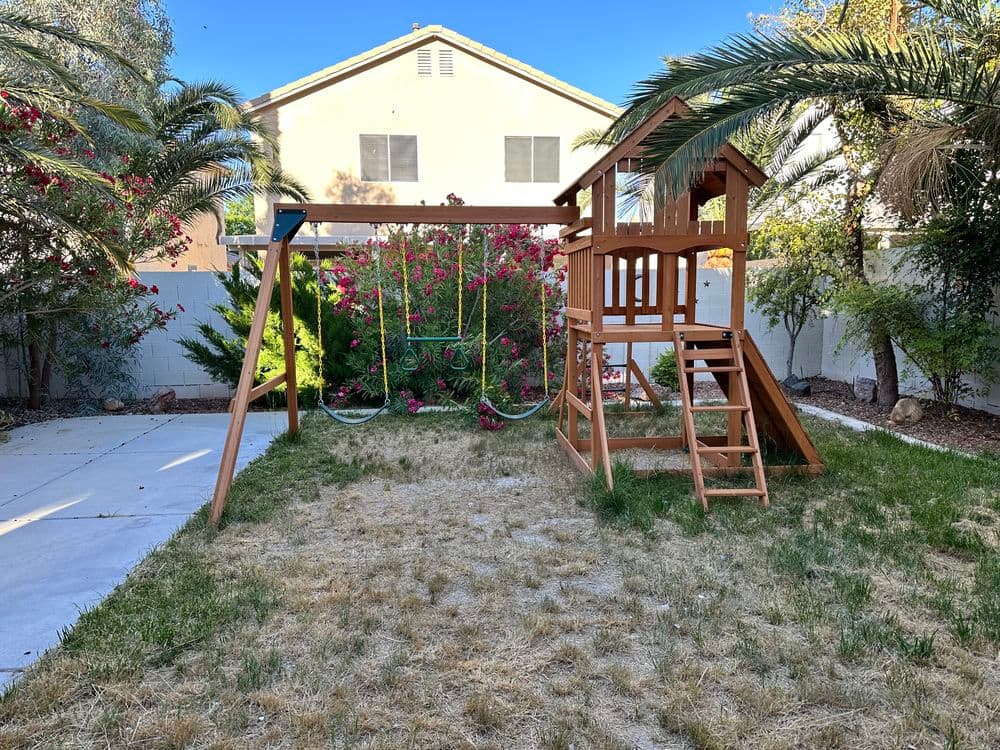 Wooden playground with swings and slide in backyard surrounded by palm trees and flowers.